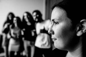 Ritratto fotografico di profilo di una persona con espressione triste o neutra, in bianco e nero, mentre in sottofondo sfocato si intravede un gruppo di persone che ridono allegramente. 35mm prime lens, depth of field, black and white film, a simboleggiare la disconnessione emotiva e la percezione alterata della risata nella depressione.
