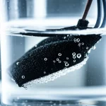 Primo piano di un elettrodo tessile flessibile nero parzialmente immerso in acqua limpida all'interno di una cella elettrolitica trasparente, macro lens 105mm, alta definizione, illuminazione laterale controllata che evidenzia la texture del tessuto e le bollicine di gas dell'elettrolisi, concetto di innovazione nel trattamento delle acque.