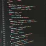 Primo piano di un monitor che mostra righe di codice software complesso con commenti di revisione evidenziati. Luce soffusa da ufficio, effetto profondità di campo. Lente prime 50mm.