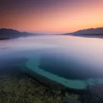 Panoramica aerea del Lago Bafa al tramonto, Turchia. Il lago salmastro riflette i colori caldi arancioni e viola del cielo. Si distinguono le aree più profonde di colore blu scuro e quelle più basse e costiere con canneti di colore verde-bruno. Montagne circondano parzialmente il lago. Obiettivo grandangolare 20mm, lunga esposizione per nuvole morbide e effetto seta sull'acqua, messa a fuoco nitida sull'orizzonte e sulla linea costiera.