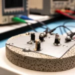 Primo piano fotorealistico di un campione di ceramica di titanato di calcio ad alta entropia (NSCST) su un banco di laboratorio, con elettrodi d'argento applicati sulla superficie lucida. Accanto, strumenti di misura elettronici parzialmente visibili e sfocati sullo sfondo. Obiettivo macro 85mm, illuminazione da laboratorio diffusa ma controllata, alta definizione sulla texture porosa ma compatta della ceramica e sulla brillantezza degli elettrodi.