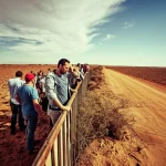 Immagine fotorealistica potente che simboleggia il 'Grande Divario': da un lato, una terra arida con persone che guardano verso un orizzonte più prospero e tecnologicamente avanzato (Nord) visibile in lontananza, separate da una barriera naturale o simbolica. Lente 35mm, stile documentaristico, colori a contrasto (caldi/desaturati vs freddi/vividi).