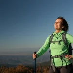 Donna matura sorridente in cima a una montagna con panorama mozzafiato, indossa abbigliamento tecnico, luce solare diretta, obiettivo 24mm, stile fotografia di viaggio realistica.