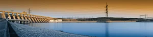 Immagine panoramica di una centrale idroelettrica moderna connessa alla rete elettrica al tramonto, con linee di trasmissione visibili che si estendono verso l'orizzonte, obiettivo grandangolare 20mm, lunga esposizione per acqua liscia nel bacino a valle, colori caldi e saturi, messa a fuoco nitida su tutta la scena.