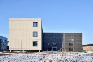 Foto grandangolare, obiettivo 18mm, che mostra la facciata di un moderno edificio sostenibile parzialmente costruito con pannelli di calcestruzzo riciclato, ambientato su uno sfondo che suggerisce un clima freddo (es. terreno leggermente innevato o cielo azzurro nitido), enfatizzando l'innovazione e la durabilità nelle costruzioni, messa a fuoco nitida, illuminazione naturale.