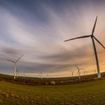 Immagine grandangolare di un paesaggio con turbine eoliche al tramonto, wide-angle 15mm, lunga esposizione per nuvole mosse, colori caldi e speranzosi, simboleggia le soluzioni e la speranza ('There's hope') nel contesto della comprensione delle verità chiave sul cambiamento climatico.