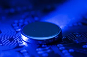 Macro fotografia di una batteria a bottone al sodio illuminata da un fascio di luce bluastra, posata su una superficie high-tech con circuiti visibili sullo sfondo. Obiettivo macro 100mm, illuminazione controllata, alta definizione.
