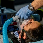 Paramedico HEMS che applica con due mani una maschera di ventilazione non invasiva (NIV) a un paziente su una barella in un ambiente esterno sfocato (es. scena di incidente stradale). Focus sulla maschera trasparente ben aderente al viso e sul tubo corrugato collegato a un ventilatore portatile visibile parzialmente. Obiettivo prime 35mm, profondità di campo ridotta, illuminazione naturale ma drammatica da scena d'emergenza.