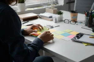 Una persona vista da dietro, seduta a una scrivania, che riorganizza attivamente post-it colorati e oggetti sul suo spazio di lavoro, simboleggiando il processo di job crafting. Luce naturale e ambiente d'ufficio moderno e accogliente. Prime lens, 35mm, depth of field.