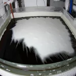 Fotografia grandangolare di una vasca sperimentale in vetro illuminata all'interno di una stanza fredda con pareti ghiacciate. All'interno della vasca, si vede acqua scura con una superficie parzialmente coperta da materiale isolante bianco (simulazione pack) e un'area aperta dove si sta formando ghiaccio sottile (simulazione polynya). Deboli scie biancastre (salamoia densa) scendono da sotto l'area ghiacciata. Obiettivo grandangolare 24mm, illuminazione fredda e drammatica, messa a fuoco nitida sull'intera scena.