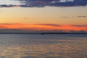 Fotografia paesaggistica della Baia di Lubecca al tramonto, obiettivo grandangolare 15mm, luce calda e dorata che si riflette sull'acqua calma, cielo con nuvole soffuse colorate di arancio e viola, una piccola barca da ricerca visibile in lontananza sulla linea dell'orizzonte, messa a fuoco nitida su tutta la scena.