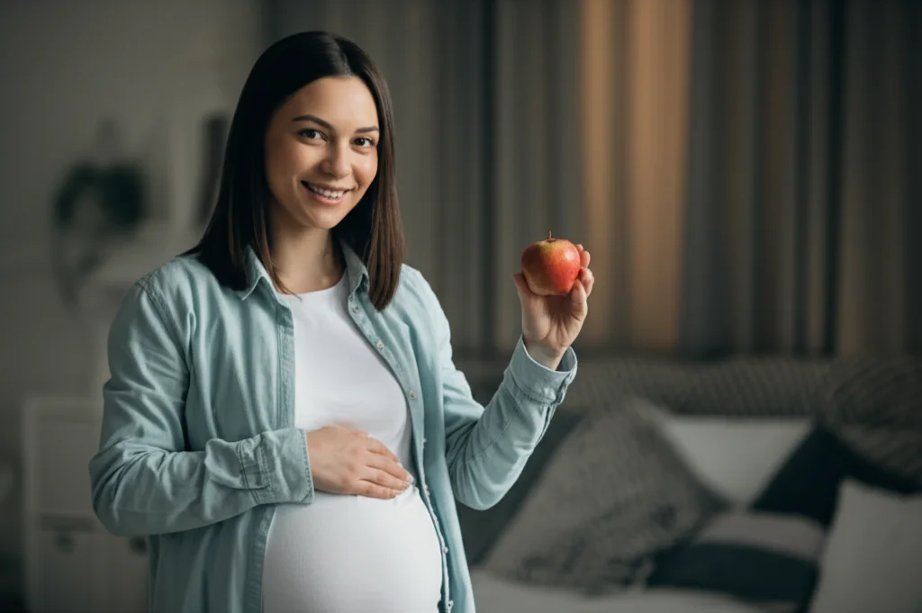 Donna incinta sorridente che tiene in mano sia una mela (simbolo di salute/peso) sia un piccolo modello della ghiandola tiroidea, guardando verso la telecamera. Ritratto 35mm, luce naturale morbida, sfondo leggermente sfocato, colori caldi.