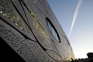 Fotografia architettonica di un edificio iconico contemporaneo con una facciata perforata dal design parametrico complesso e organico. La luce del tardo pomeriggio filtra attraverso le perforazioni creando pattern luminosi all'interno visibili da fuori. Obiettivo grandangolare 24mm, profondità di campo, alta definizione, dettagli nitidi della texture del materiale della facciata (metallo o cemento composito).