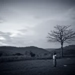 Immagine suggestiva e rispettosa di un paesaggio rurale dell'Uganda Orientale al crepuscolo, con una figura femminile solitaria e indistinta in lontananza vicino a un albero spoglio. Obiettivo grandangolare 24mm, lunga esposizione per cieli sfumati e nuvole morbide, colori desaturati tendenti al blu e grigio duotone, focus nitido sul paesaggio, evocando un senso di mistero, isolamento e tristezza legati alle pratiche sui nati morti.