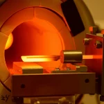 Immagine fotorealistica di un setup sperimentale per la separazione a secco del Radioscandio: un tubo di quarzo sotto vuoto spinto inserito in una fornace tubolare. All'interno, un sottile foglio di titanio (circa 50 micron) è incandescente (1100°C), emettendo una luce arancione intensa. Di fronte al foglio, un 'dito freddo' metallico (potrebbe essere rivestito d'oro) è posizionato per catturare gli atomi di Scandio-47 evaporati. Macro lens, 90mm, high detail, precise focusing, controlled lighting per enfatizzare il contrasto tra il caldo e il freddo e l'ambiente high-tech.