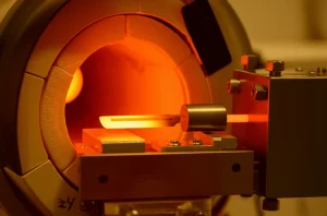 Immagine fotorealistica di un setup sperimentale per la separazione a secco del Radioscandio: un tubo di quarzo sotto vuoto spinto inserito in una fornace tubolare. All'interno, un sottile foglio di titanio (circa 50 micron) è incandescente (1100°C), emettendo una luce arancione intensa. Di fronte al foglio, un 'dito freddo' metallico (potrebbe essere rivestito d'oro) è posizionato per catturare gli atomi di Scandio-47 evaporati. Macro lens, 90mm, high detail, precise focusing, controlled lighting per enfatizzare il contrasto tra il caldo e il freddo e l'ambiente high-tech.