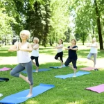 Gruppo diversificato di donne di mezza età che partecipano a una lezione di yoga dolce all'aperto in un parco rigoglioso, guidate da un'istruttrice. Alcune sono in piedi, altre sedute su tappetini. Espressioni concentrate ma serene. Obiettivo grandangolare 24mm per includere il gruppo e l'ambiente naturale. Luce solare filtrata dagli alberi.