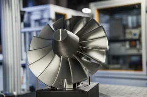 Primo piano fotorealistico di una pala di turbina complessa realizzata in lega Mo-Si-B grigio metallico lucido, appena stampata in 3D con tecnologia PBF-EB. La pala mostra dettagli fini e superfici lisce. L'ambiente circostante è un laboratorio high-tech pulito ma leggermente sfocato sullo sfondo. Obiettivo prime 35mm, profondità di campo ridotta per isolare la pala, illuminazione drammatica laterale che evidenzia la forma complessa e la finitura metallica brillante.