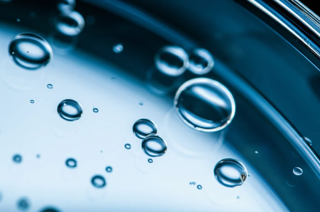 Primo piano macro di microgoccioline sferiche trasparenti (MMD di polilattato) che fluttuano in una soluzione acquosa all'interno di una piastra di Petri. Illuminazione laterale controllata che evidenzia la tridimensionalità delle gocce. Obiettivo macro 105mm, alta definizione, sfondo leggermente sfocato per concentrare l'attenzione sulle goccioline, simulando protocellule primitive in un ambiente di laboratorio.