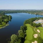 Veduta aerea del Lago Berendonck, un lago urbano nei Paesi Bassi, circondato da alberi decidui, un campo da golf su un lato e strutture ricreative sull'altro. Obiettivo grandangolare 15mm per catturare l'estensione del lago (0.45 km2) e l'ambiente circostante urbanizzato ma verde. Luce diurna morbida, sharp focus, acqua calma che riflette parzialmente il cielo.