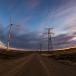 Fotografia wide-angle, 15mm, di un parco eolico in una zona rurale remota al tramonto, con lunghe linee elettriche che si estendono verso l'orizzonte, simboleggiando l'integrazione in reti deboli. Esposizione lunga per nuvole setose, messa a fuoco nitida.