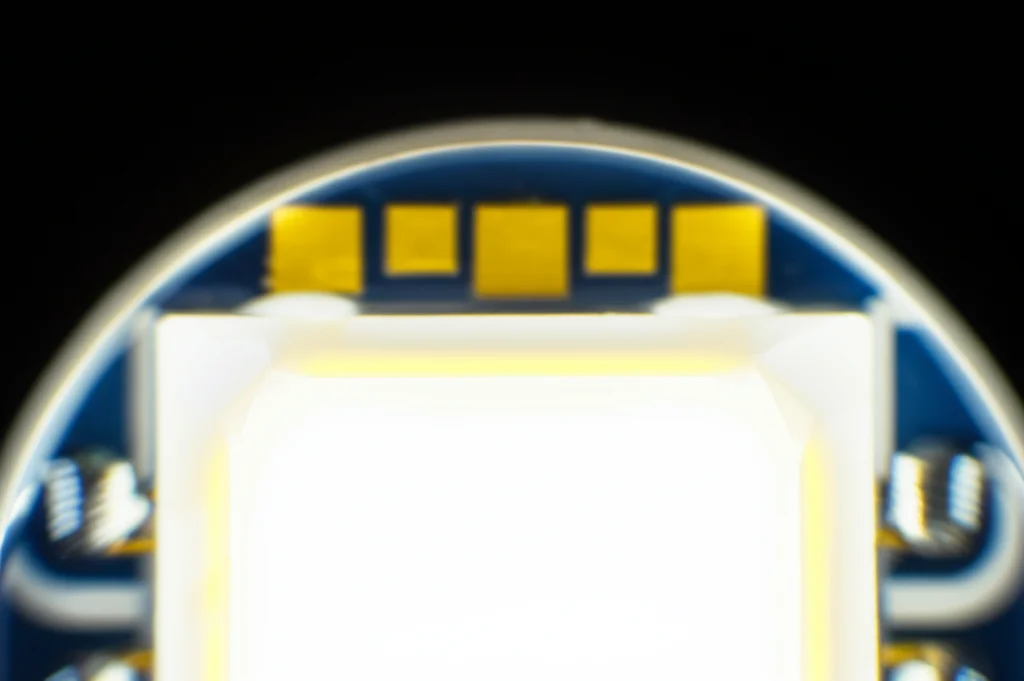 Immagine macro ad alta definizione di un moderno chip LED bianco acceso, obiettivo 100mm, che mostra i dettagli interni come i fosfori gialli e i sottili fili di collegamento dorati, illuminazione da studio controllata per massima nitidezza e dettaglio, sfondo scuro per contrasto.