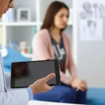 Medico specialista in gastroenterologia che esamina attentamente i risultati del monitoraggio terapeutico di un farmaco (TDM) su un tablet, con un paziente sullo sfondo sfocato in un ambulatorio moderno. Lente prime 50mm, profondità di campo, illuminazione naturale controllata.