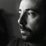 Ritratto di un adulto autistico che guarda con speranza verso una fonte di luce laterale, simboleggiando il miglioramento emotivo post-DBT. Obiettivo prime 35mm, luce soffusa e calda, profondità di campo ridotta per focalizzare sullo sguardo. Bianco e nero, film noir style.