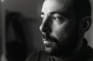 Ritratto di un adulto autistico che guarda con speranza verso una fonte di luce laterale, simboleggiando il miglioramento emotivo post-DBT. Obiettivo prime 35mm, luce soffusa e calda, profondità di campo ridotta per focalizzare sullo sguardo. Bianco e nero, film noir style.