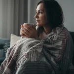 Fotografia stile reportage, una donna seduta comodamente su un divano, avvolta in una coperta morbida, tiene una tazza calda tra le mani, guarda fuori dalla finestra con un'espressione serena ma consapevole, luce naturale morbida, 35mm, profondità di campo, colori tenui, rappresentando il concetto di autogestione e benessere a casa per l'endometriosi.