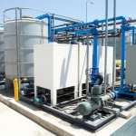 Impianto pilota di trattamento delle acque reflue tramite elettrocoagulazione, visualizzato con obiettivo grandangolare 20mm per catturare l'intera scena con messa a fuoco nitida sotto luce diurna. Si vedono serbatoi cilindrici e rettangolari collegati da tubazioni in un ambiente industriale pulito.