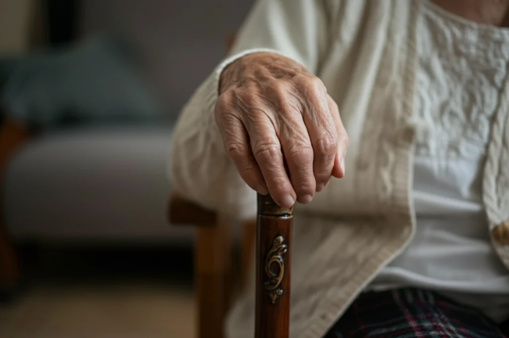Primo piano di una mano anziana che si appoggia con forza su un bastone da passeggio decorato, simbolo della fragilità ma anche della determinazione e resilienza dopo una frattura d'anca. Luce laterale morbida che evidenzia le rughe e la texture della pelle e del legno, obiettivo prime 50mm, profondità di campo ridotta per sfocare lo sfondo.