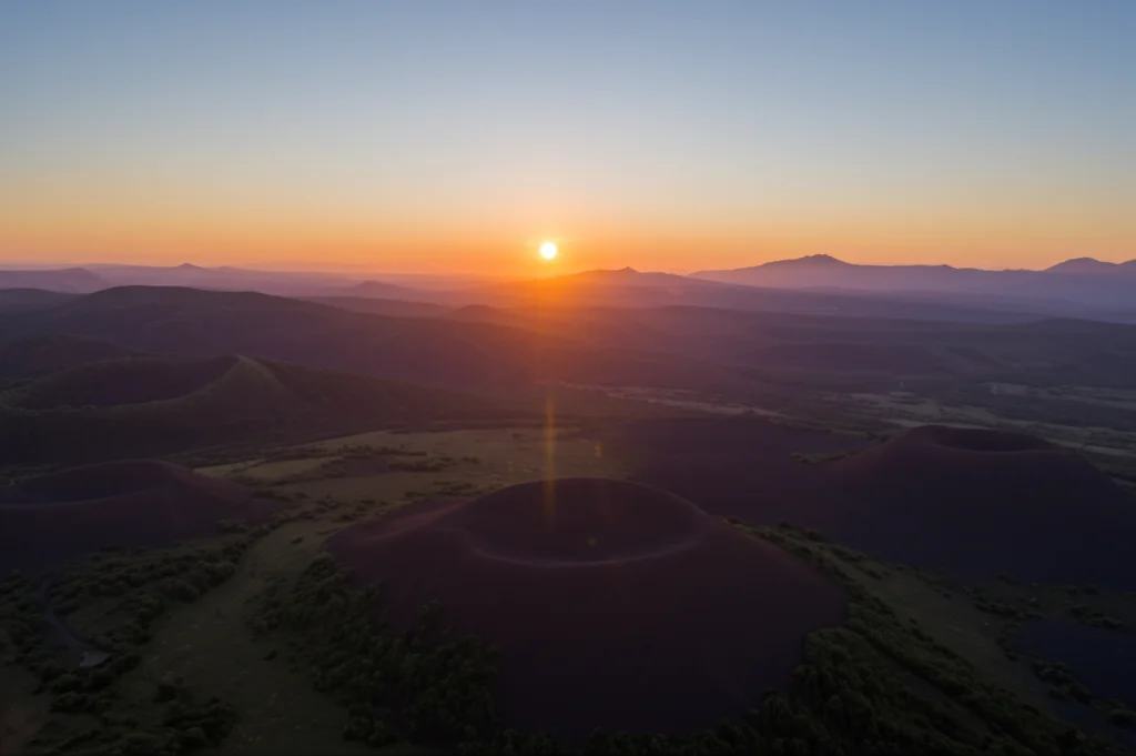 Veduta aerea panoramica del Campo Vulcanico di Perșani, Romania, al tramonto. Si distinguono le forme coniche dei coni di scorie (come Hegheș) e le aree delle cave (come Racoș). Luce calda radente che esalta le forme del rilievo. Obiettivo grandangolare 24mm, lunga esposizione, messa a fuoco nitida.