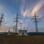 Immagine fotorealistica di una moderna rete di distribuzione elettrica al tramonto, wide-angle 20mm, con linee elettriche che si intersecano con campi di pannelli solari (PV-DG) e un piccolo edificio tecnologico che rappresenta un DSTATCOM, long exposure per nuvole sfumate, sharp focus.
