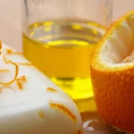 Immagine fotorealistica, obiettivo macro 100mm, che mostra una saponetta dall'aspetto naturale infusa con minuscole pagliuzze di buccia d'agrume appoggiata accanto a un piccolo becher di olio da cucina esausto dorato e una buccia d'arancia fresca, alto dettaglio, messa a fuoco precisa, illuminazione calda controllata.