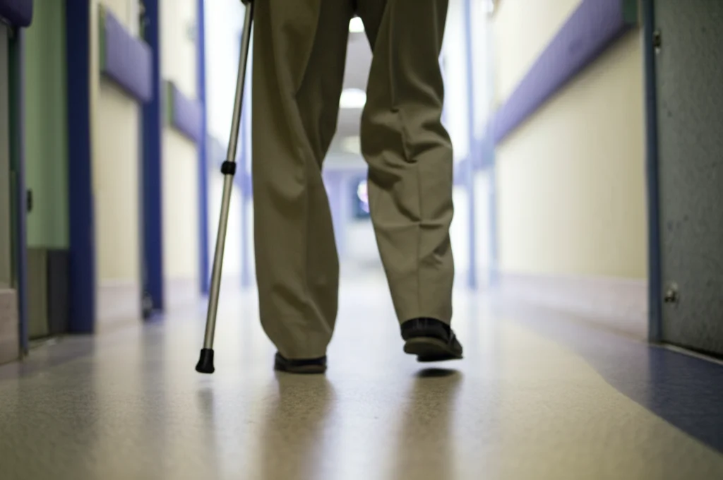 Fotografia di un paziente anziano, visto dai fianchi in giù, che cammina lentamente lungo un corridoio d'ospedale luminoso ma tranquillo. Focus sui piedi e sul pavimento. Obiettivo 35mm, profondità di campo ridotta per sfocare leggermente lo sfondo. Toni neutri.