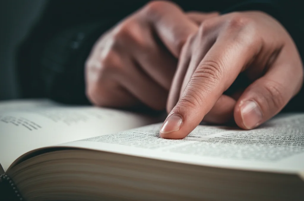 Primo piano macro di una mano di insegnante che indica un punto specifico su un libro aperto tenuto da uno studente, simboleggiando l'attenzione al dettaglio nell'insegnamento. Obiettivo macro 100mm, illuminazione laterale morbida, alto dettaglio sulla texture della pagina e delle dita, messa a fuoco selettiva sul punto di contatto.