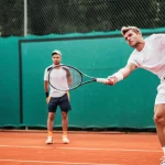 Tennista professionista durante una sessione di screening funzionale sul campo da tennis in terra battuta, supervisionato attentamente da un preparatore atletico. Fotografia sportiva d'azione, teleobiettivo 200mm, alta velocità dell'otturatore per catturare il movimento, profondità di campo ridotta per enfatizzare l'atleta.