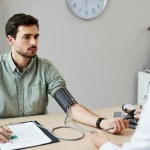 Giovane adulto (circa 25 anni) durante una misurazione della pressione arteriosa in un ambiente medico moderno. L'espressione è neutra ma attenta, suggerendo consapevolezza sulla propria salute. Fotografia ritratto, obiettivo 35mm, luce naturale laterale, profondità di campo media per includere dettagli dello sfigmomanometro, stile fotorealistico.