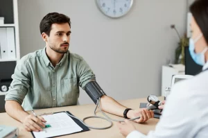 Giovane adulto (circa 25 anni) durante una misurazione della pressione arteriosa in un ambiente medico moderno. L'espressione è neutra ma attenta, suggerendo consapevolezza sulla propria salute. Fotografia ritratto, obiettivo 35mm, luce naturale laterale, profondità di campo media per includere dettagli dello sfigmomanometro, stile fotorealistico.