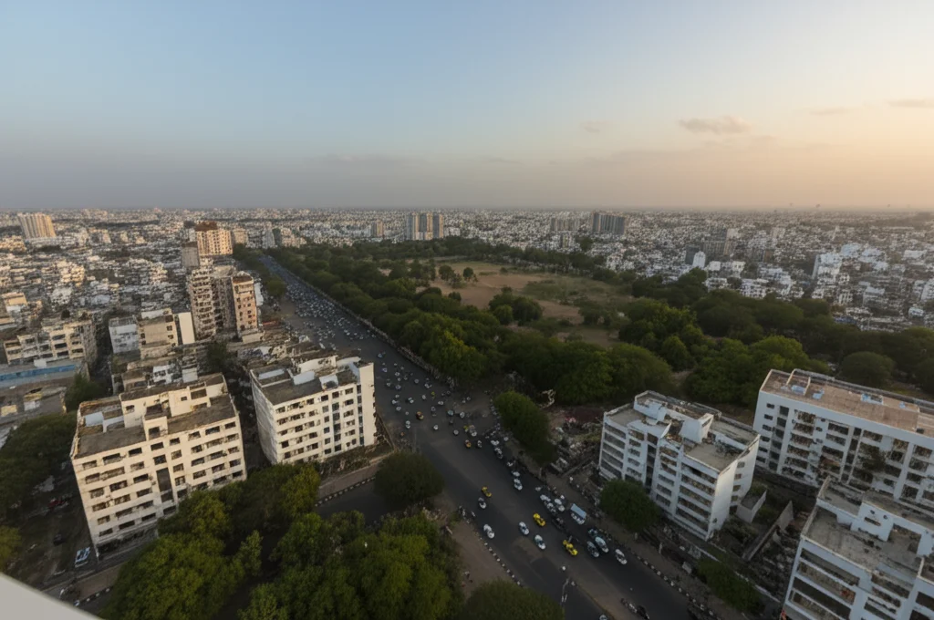 Veduta aerea di una città indiana di medie dimensioni al tramonto, che mostra un mix di edifici bassi e alti, strade trafficate con veicoli e alcune macchie significative di verde (parchi o viali alberati). Fotografia paesaggistica, obiettivo grandangolare 20mm, luce calda del tramonto che crea lunghe ombre, alta definizione, focus nitido su tutta la scena.