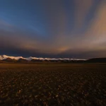 Fotografia paesaggistica grandangolare, 15mm, di campi coltivati sull'altopiano tibetano al tramonto, con il terreno scuro e fertile in primo piano e le montagne illuminate sullo sfondo. Messa a fuoco nitida, colori caldi, lunga esposizione per cielo suggestivo.