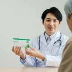 Immagine fotorealistica di un medico giapponese sorridente che mostra una scatola di farmaci SGLT2i a un paziente anziano in un ambiente clinico luminoso e moderno, profondità di campo, lente prime 50mm, per rappresentare l'applicazione clinica e i benefici per i pazienti.