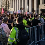 Immagine fotorealistica di una folla divisa da una barricata della polizia, da un lato manifestanti pacifici con cartelli contro l'odio, dall'altro lato sostenitori di un evento controverso che entrano in un edificio. Scena urbana al tramonto, luce drammatica, obiettivo zoom 24-70mm, stile fotogiornalistico.