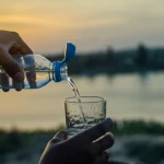 Immagine fotorealistica, obiettivo 35mm, che mostra una mano che versa acqua da una borraccia in un bicchiere trasparente, con sullo sfondo sfocato un paesaggio fluviale del Ghana al tramonto, duotone blu e ocra, profondità di campo per evidenziare l'acqua.