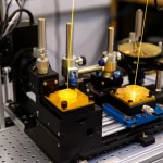 Immagine fotorealistica di un setup da laboratorio ottico complesso con laser Terahertz. Un interferometro di Mach-Zehnder è al centro, con specchi dorati e polarizzatori a griglia metallica che dividono e ricombinano un fascio THz visibile (rappresentato artisticamente come un'onda traslucida). Luce ambientale controllata, focus preciso sui componenti chiave come lo stage di traslazione micrometrica, obiettivo macro 85mm, alta definizione, profondità di campo ridotta per enfatizzare l'interferometro.