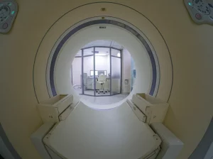 Interno di una sala di Risonanza Magnetica clinica moderna, vista grandangolare 24mm. Il tunnel dello scanner è illuminato centralmente, con la console di controllo visibile in secondo piano attraverso la finestra schermata. Atmosfera high-tech e pulita, focus nitido sull'attrezzatura RM.