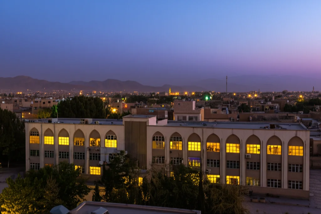 Veduta urbana di Herat, Afghanistan, al tramonto, con focus su un edificio sanitario riconoscibile ma integrato nel contesto cittadino. Obiettivo grandangolare 24mm, esposizione lunga per catturare le luci della città, messa a fuoco nitida sull'architettura e sulla vita circostante.