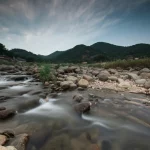 Paesaggio coreano con affioramenti di granito fratturato e un ruscello che scorre in primo piano, cielo parzialmente nuvoloso. Landscape wide angle 15mm, luce naturale morbida del tardo pomeriggio, sharp focus, acqua del ruscello leggermente mossa (smooth water).