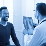 Immagine fotorealistica di un paziente etiope sorridente che riceve i risultati di una radiografia da un operatore sanitario in un ospedale pubblico. L'ambiente è luminoso e moderno, simboleggiando l'impatto positivo della teleradiologia. Ritratto con obiettivo 35mm, luce calda, effetto duotone blu e bianco per un tocco emotivo.